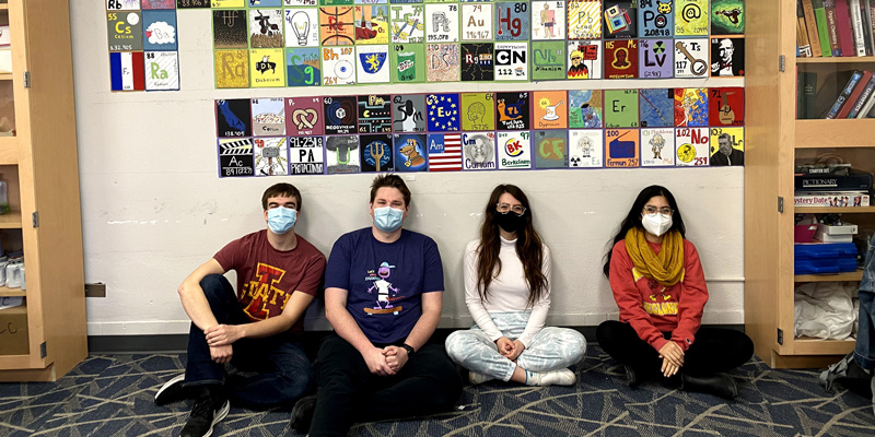 Students wearing masks, sitting beneath a decorative periodic table