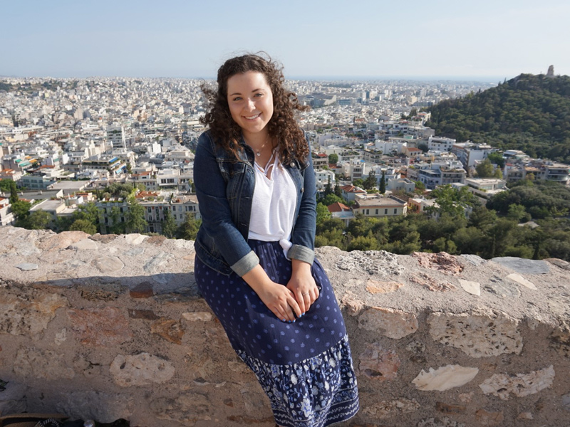 Sophie Catus sitting on a brick wall with view of a European city in the background